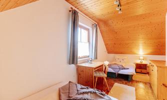A cozy bedroom with wooden walls and a sloped roof. There are two beds, a table, and plenty of natural light coming in through a window.