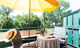 A cozy balcony with a yellow sunshade and a small table. On the table lie a book and sunglasses, surrounded by green trees.