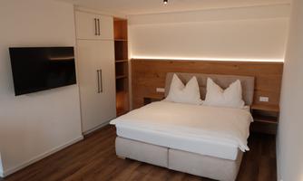 A modern bedroom with a large bed and two pillows. A television hangs on the wall, and there is a wooden wardrobe as well as indirect lighting.
