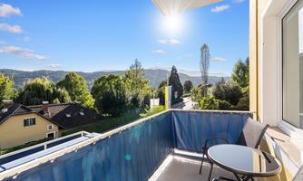 A sunny balcony with a table and two chairs. In the background, green hills and a clear blue sky can be seen.