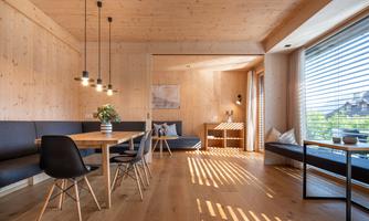 A modern living room with light wood paneling and large windows. The space is furnished with a dining table, cozy seating, and decorative elements.