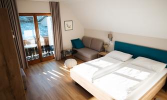 A modern room with a large double bed and a cozy sofa. The windows offer a view of a balcony and let in plenty of natural light.