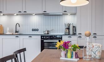 A modern kitchen with white cabinets and a wooden dining table. A decorative flower vase is on the table, and various kitchen utensils are visible.