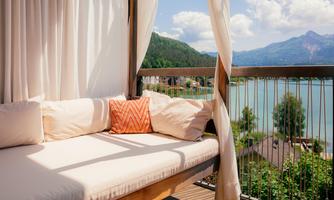 A comfortable lounge bed on a balcony overlooking a lake and mountains. The atmosphere is inviting and relaxed.