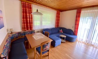 A cozy living room with a blue couch and a wooden dining area. Large windows provide plenty of daylight and a friendly atmosphere.