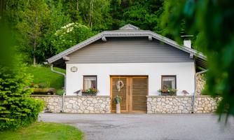 A beautiful house with a bright stone foundation and a gray roof. Flowers adorn the windows, and the surroundings are green and inviting.