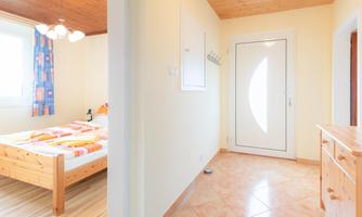 A bright bedroom with a double bed and colorful blankets. The entrance area has a white door and wooden furniture.
