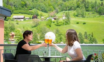 Two women are toasting with drinks and enjoying the view of a green landscape. In the background, huts and trees can be seen.