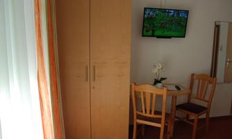 A simple room with a wooden cupboard, a table, and two chairs. A television is hanging on the wall and a small mirror is visible.