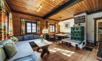 A cozy living room with wood paneling and large windows. There is a seating area, a table, and a traditional stove.