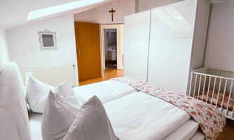 A bright bedroom with a large bed and two pillows. In the background, there is a wardrobe and a children's bed.