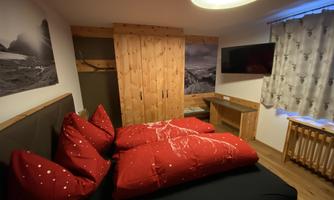 A cozy bedroom with a large bed and red pillows. The walls are bright with wooden details and a television as well as windows with curtains.