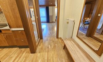 An inviting hallway with wooden flooring and a small seating area. In the background, a bright room with a window can be seen.