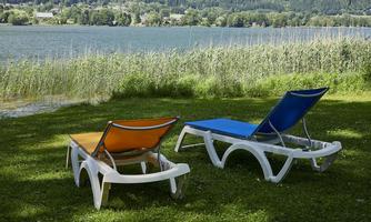 Two loungers are placed on green grass by the shore of a body of water. In the background, trees and gentle hills can be seen.