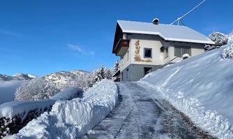 A large, white house on a snow-covered slope. The sky is clear and blue, while trees and paths are covered with fresh snow.