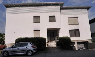 A plain white residential building with several windows. In front of the building is a gray car.