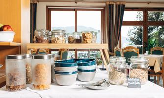 A buffet with various cereals and dried fruits in jars and bowls. In the background, you can see a bright room with windows and cozy seating areas.