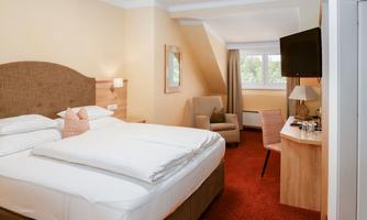 An elegant hotel room with a large bed and cozy furniture. The room has a television, a desk, and a window with a view to the outside.