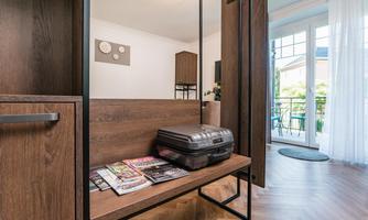 A modern room with a wooden shelf and a suitcase on a bench. Large windows offer a view of the balcony and the outside area.