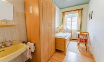A simple room with a single bed, a wooden wardrobe, and a table with a chair. The room has a window with curtains and a small sink area.