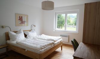A bright bedroom with a double bed and two nightstands. Large windows offer a view of greenery and provide plenty of daylight.