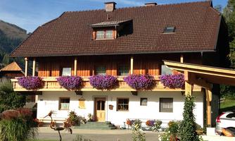 A traditional alpine house with wooden cladding. It has flower boxes with purple flowers and a beautiful garden in front of the door.