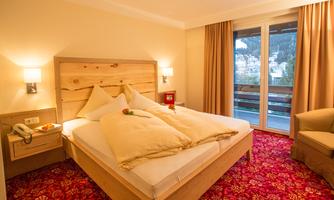 A cozy bedroom with a large bed, surrounded by light wood and soft pillows. From large windows, one looks out at the mountains, and the living room is stylishly furnished.