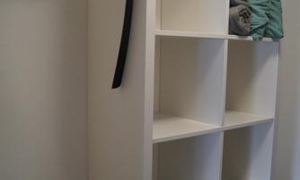 A simple, white wardrobe with open compartments. On the left side, there are hangers and a folded blanket is on top.