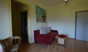 A cozy living room with a red sofa and a small table. A colorful painting hangs on the wall, and there is a wooden stool in the corner.