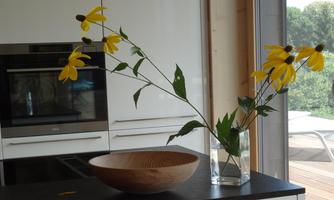 An elegant kitchen ambiance with a wooden bowl and yellow flowers in a vase. In the background, you can see modern kitchen equipment and plenty of daylight.