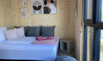 A cozy room with a bed, decorative pillows, and modern artworks on the wall. Large windows provide plenty of natural light and an inviting atmosphere.