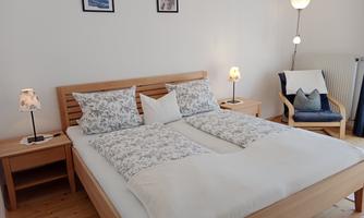 A cozy bedroom with a large double bed and decorative pillows. Next to the bed are nightstands with table lamps and an armchair in the corner.