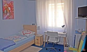 A bright, modern room with a bed, a desk, and a television. The windows are covered with curtains and there is a colorful chair.