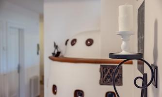 A bright room with a curved white wall decoration and a candle on a candle holder. In the background, a door and a tiled stove can be seen.