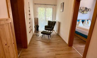 A bright hallway with wooden flooring and a green plant. At the end of the hallway, there is a comfortable armchair and a window with curtains.