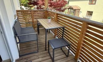 A cozy balcony with a table and two chairs. In the background, trees and various buildings can be seen.