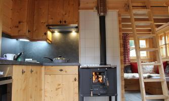 A cozy kitchen in a wooden house with a wood stove. The decor is simple and rustic, with warm wooden surfaces and a staircase in the background.