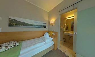 A cozy single room with a simple bed and a small table. The bathroom is accessible through a glass door and offers modern amenities.