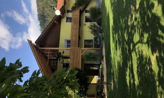 A charming house with a yellow facade and wooden balcony. The garden is green and inviting, surrounded by trees and hills.