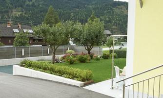 A well-kept garden landscape with trees and colorful flowers. In the background, mountains and buildings can be seen.