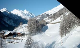 A picturesque winter landscape with snow-covered mountains and a clear blue sky. In the foreground, there are snow-covered hills and a few cozy houses.