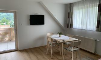 A bright living and dining room with a table and chairs. Windows with curtains offer a view of nature.