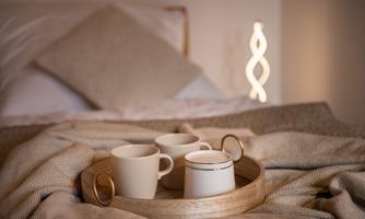 A cozy bed with a blanket and a tray on which three cups are placed. In the background, a modern lamp is glowing.