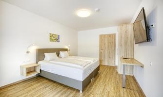 A modern hotel room with a double bed, a television, and wooden flooring. There is a simple, bright decor and a window to the door.