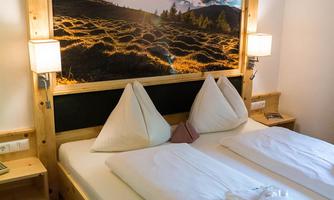 A cozy hotel room with a large bed and a decorative headboard. On the wall hangs a beautiful landscape painting featuring mountains and green meadows.