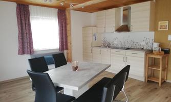 A modern kitchen with a dining table and black chairs. There are windows with curtains and a light wooden ceiling.