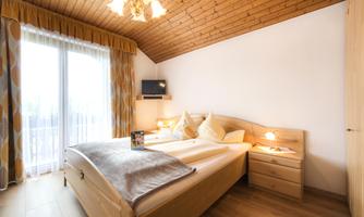 A cozy bedroom with a large bed and wooden ceiling. Large windows let in plenty of daylight.