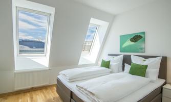 A modern bedroom with two beds and green cushions. The walls are bright and there are large windows that let in plenty of daylight.