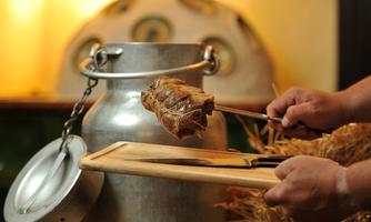 A metal container is standing on a tabletop, while someone is holding a piece of meat with a fork. In the background, some straw decorations are visible.