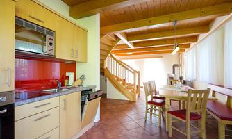 A modern kitchen with wooden shelves and red accents. In the background, there is a bright dining area with a table and a staircase.